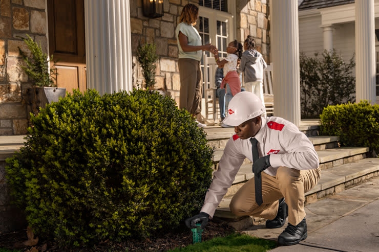 An Orkin Pro inspecting for termites in customers- front yard while they stand on their porch